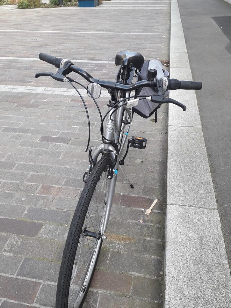 A silver ladies bike parked up next to the pavement with panniers attached.