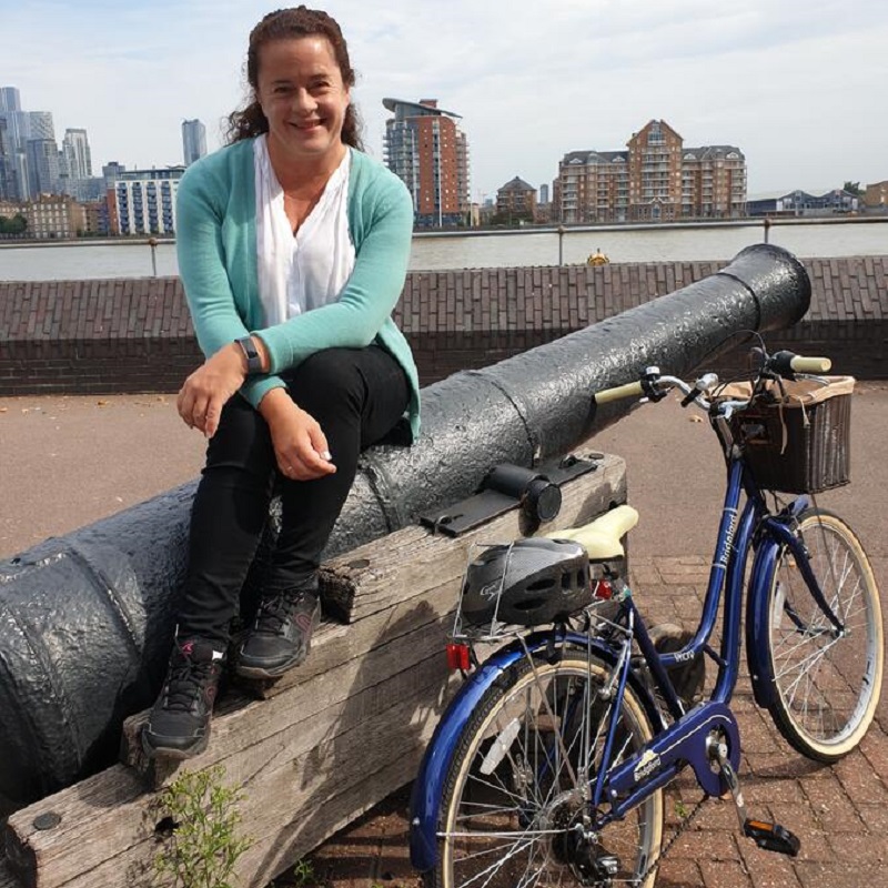 Joanna wears a teal cardigan while sitting on a cast iron cannon by the Thames, with her cream seated electric blue bike leaning next to her.