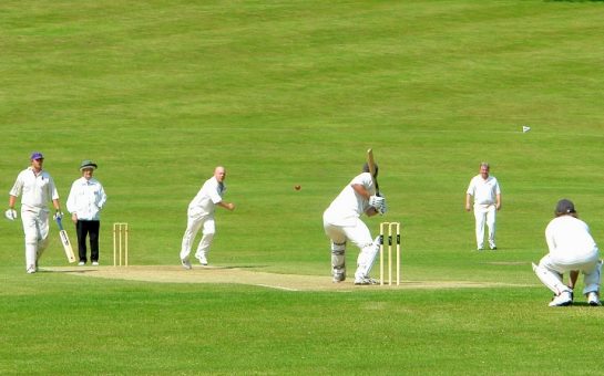 bowler delivers a full length ball pitched at off stump to a batsmen waiting to hit his shot, with a wicket keep tucked behind, in a lush green setting