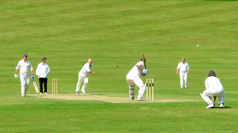 bowler delivers a full length ball pitched at off stump to a batsmen waiting to hit his shot, with a wicket keep tucked behind, in a lush green setting