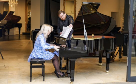 Harriet Stubbs, a woman in a glamorous blue dress, sits at a black, open grand piano. Her head is bent in concentration. A male stands to her right, turning the pages of her music on the stand.