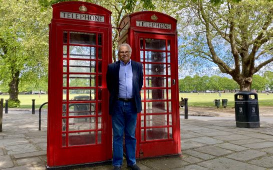 Chairman of Richmond Society, Barry May, standing in a shaded spot under a tree in front of two red English phone booths. The booths are the well known booths that are synonymous with the UK. Behind them is a park which has the sun casting on it.