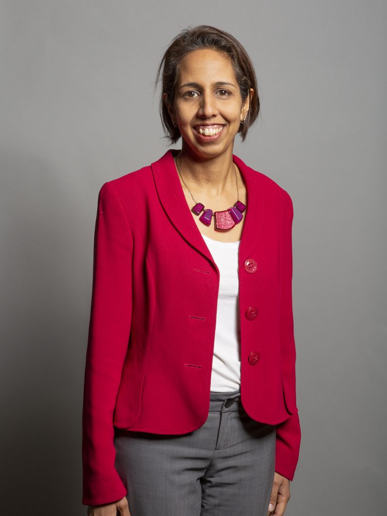 Munira Wilson portrait. She is wearing a pink jacket and grey trousers and is smiling. 