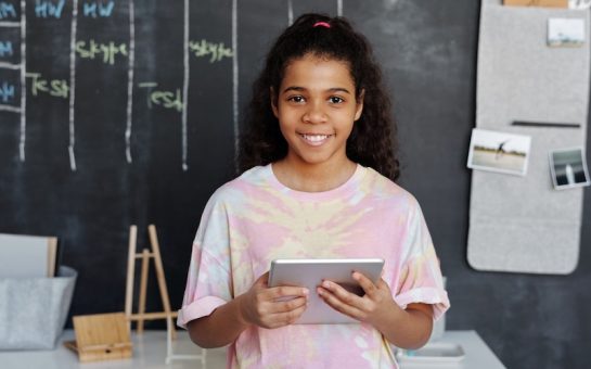 picture of a girl holding a device. Behind her is a blackboard. She is wearing a tie dye shirt.