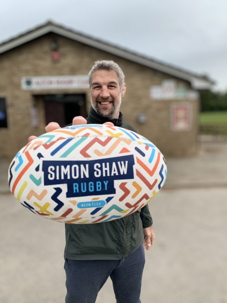 Ex-England international Simon Shaw holding a rugby ball toward the camera with Simon Shaw Rugby written on it and many colourful lines