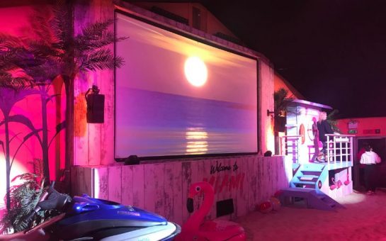 Miami Nights screen room with pink lighting, plam trees, a speed boat proper and an inflatable flamingo around the sides of the cinema screen. A sunset is on the screen.