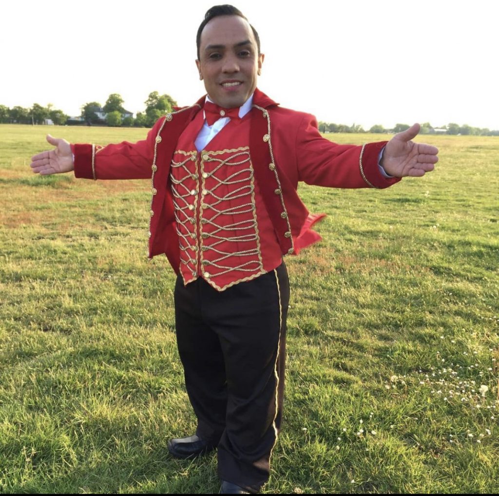 A photo of Paulo Dos Santos in a red ring master's jacket with gold detailing and buttons. His trousers are black with gold outer-lining. He is also wearing a red bow-tie. He is standing on a green field.
