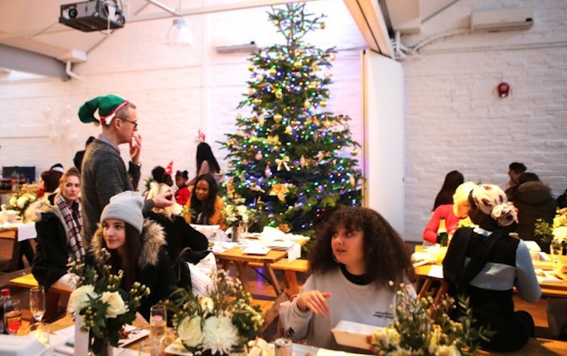 Young people sit at festively decorated tables with a Christmas tree in the background.