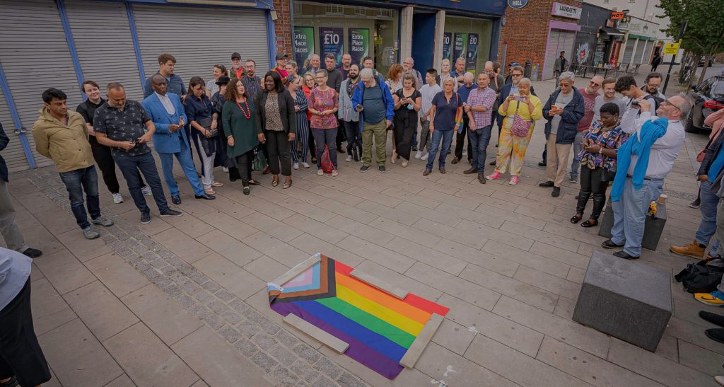 A crowd gathered around a progress pride flag covering the plaque at the unveiling ceremony