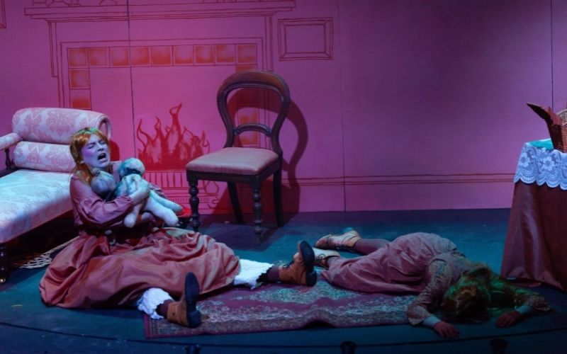 A woman in 19th-century dress and a wig cries on the floor while holding two rag dolls, and a second woman lies dead next to her
