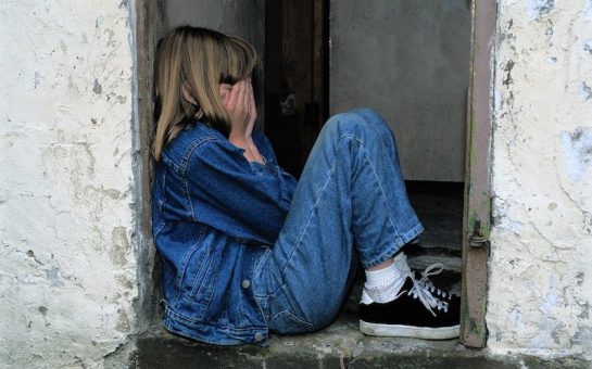 A child sitting on an outdoor step, face covered with her hands in fear