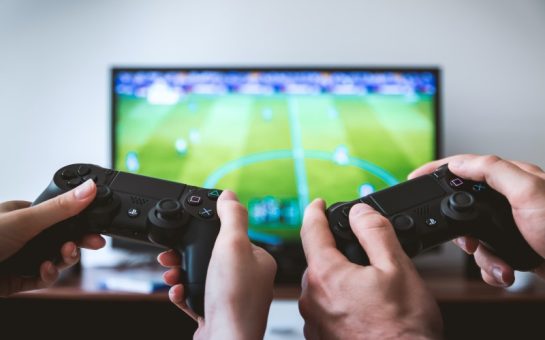 Two pairs of hands holding PlayStation controllers in front of a monitor