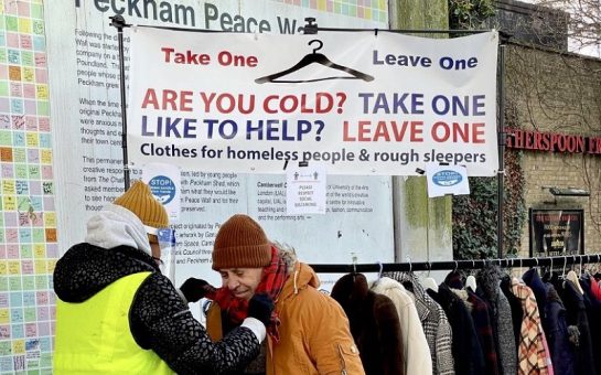 A volunteer in a high-vis jacket helps a man put on a scarf in front of a clothes rail of winter clothes. A sign above says "Take One Leave One. Are you cold? Take one. Like to help? Leave one. Clothes for homeless people and rough sleepers"