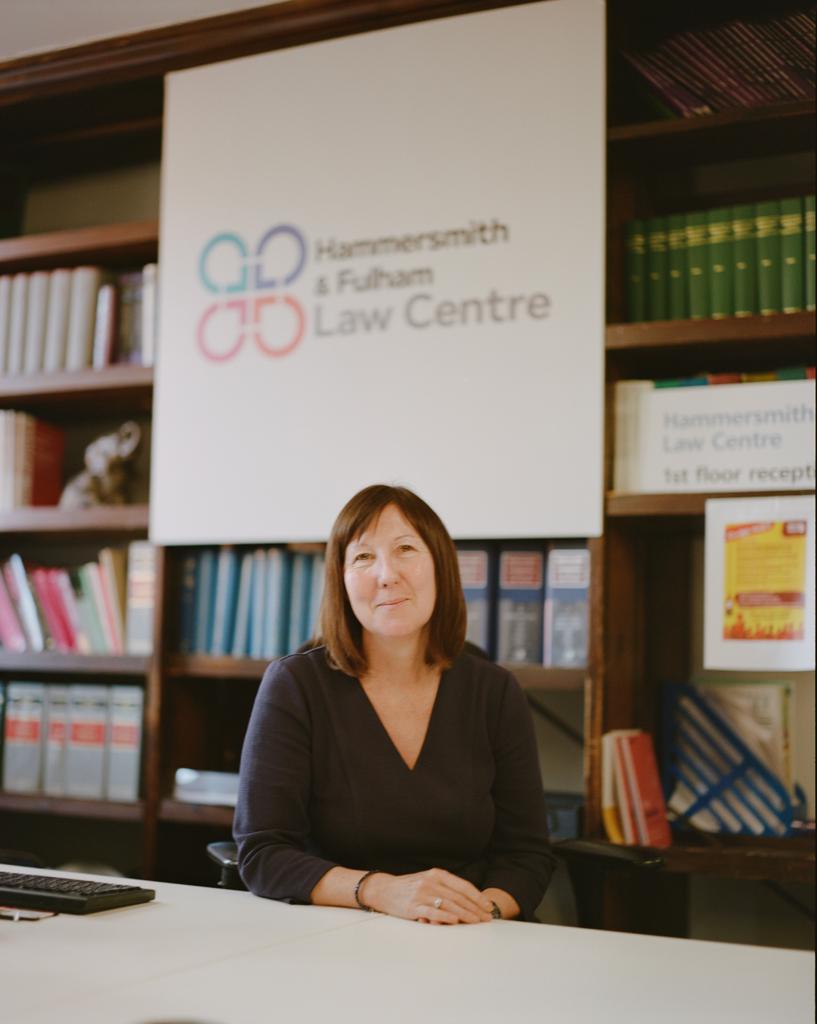 Photo of caucasian woman sitting in office with a sign behind her reading "Hammersmith and Fulham Law Centre." She is wearing a dark top.