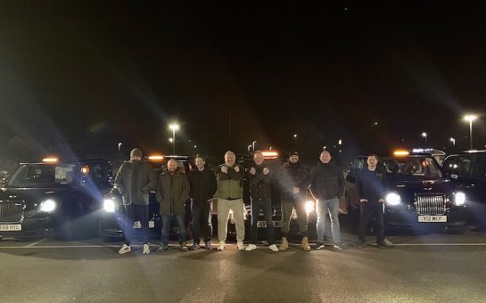 Eight of the drivers stand in front of five of the six taxis. They are in a dark carpark and the headlights are on.