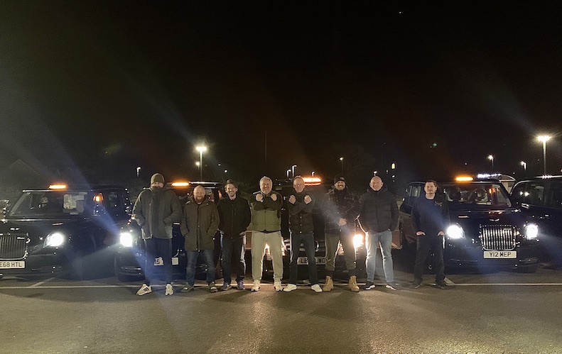 Eight of the drivers stand in front of five of the six taxis. They are in a dark carpark and the headlights are on. 