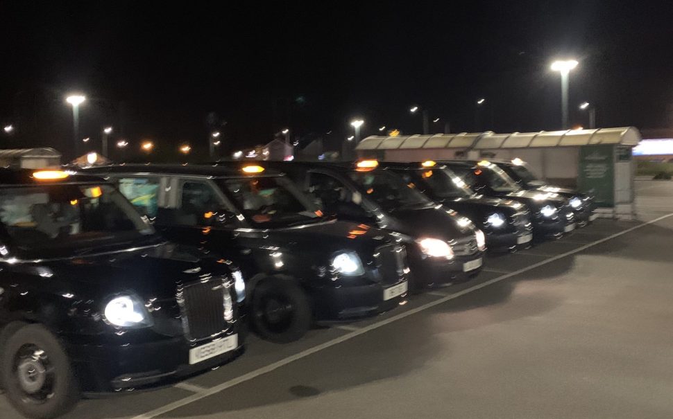 Six black taxis are parked in a line in a carpark. It is night time and blurry.