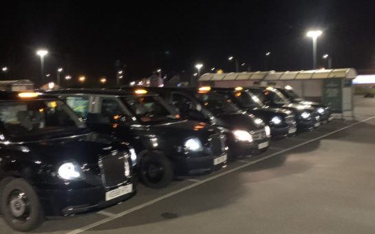 Six black taxis are parked in a line in a carpark. It is night time and blurry.