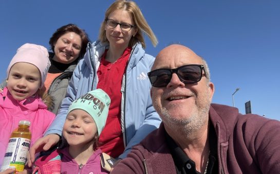 Richard Gough and the family who have fled to the UK from Kyiv smile together against a blue sky. Grandmother, mother, and two daughters stand behind Richard in a selfie.