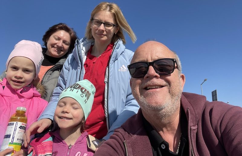 Richard Gough and the family who have fled to the UK from Kyiv smile together against a blue sky. Grandmother, mother, and two daughters stand behind Richard in a selfie.