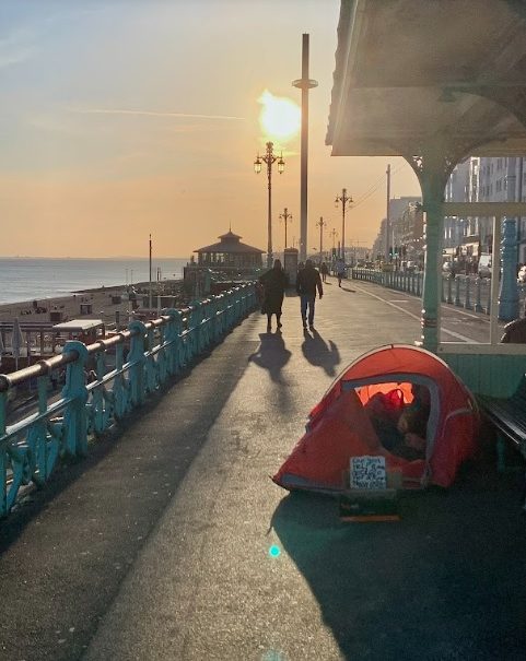 Local residents enjoy an evening stroll along the promenade as a homeless citizen sets up his tent for the night. Photo by Eve Salusbury