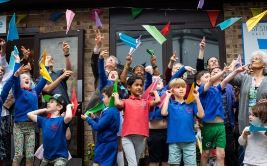 A group of primary school children and elderly people are stood in front of a building, throwing colourful paper aeroplanes into the air