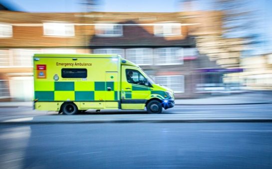 An ambulance is driving quickly down a street with houses behind. The photo is slightly blurred to show the speed.