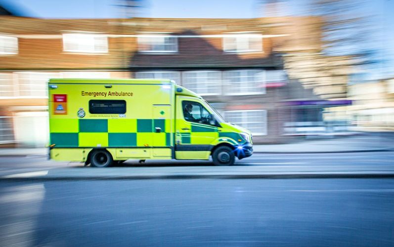 An ambulance is driving quickly down a street with houses behind. The photo is slightly blurred to show the speed.