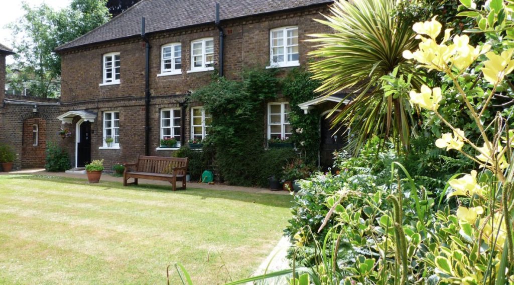 The front of Houblon's Almshouses in Richmond owned by the Richmond Charities, showing a bench, lawn and flowers with plant pots outside the housing.