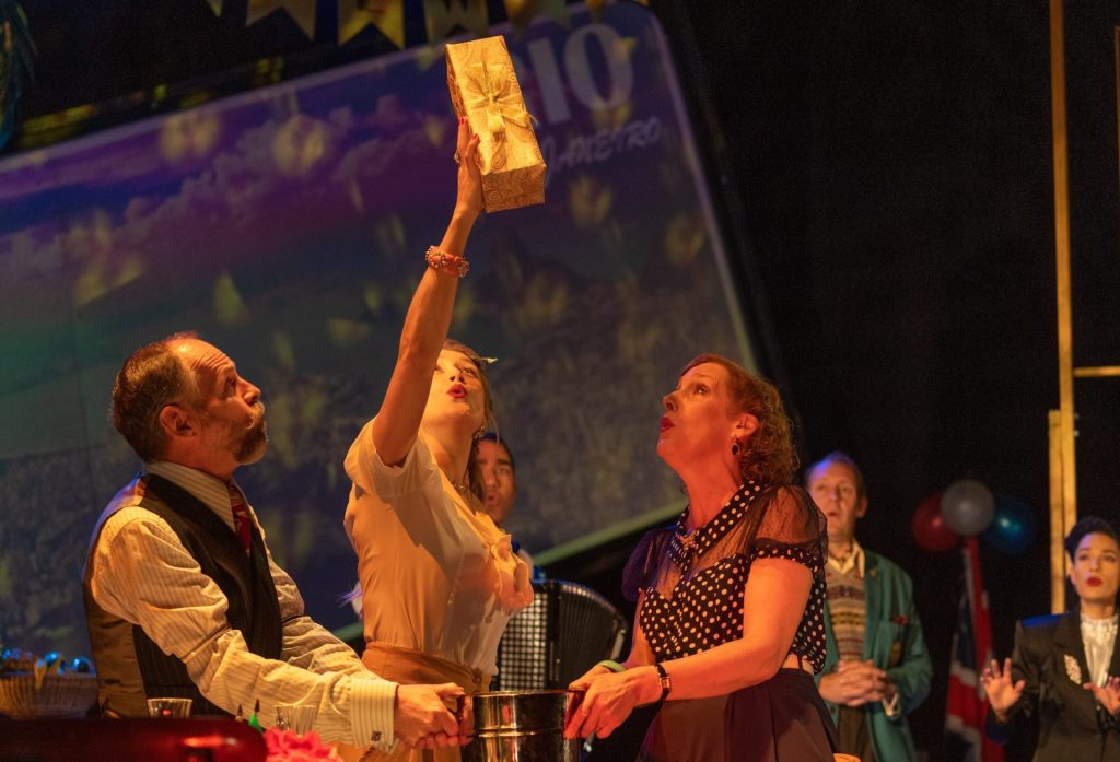 The cast of the Lavender Hill Mob on stage. They are all shocked and looking at a gold box wrapped in a ribbon. 