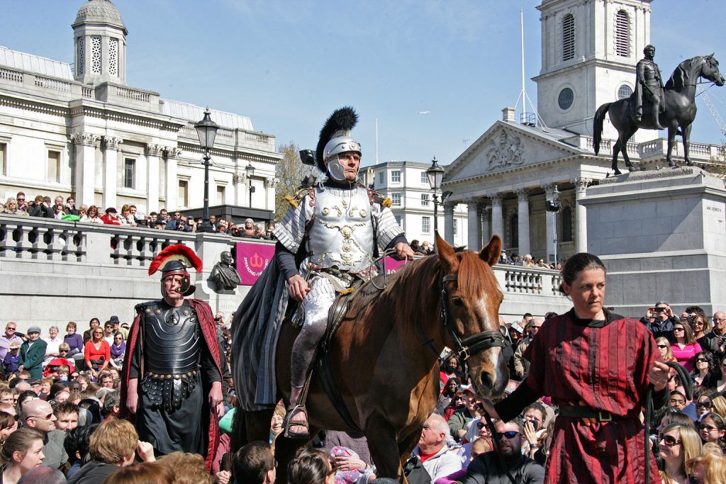 Roman soldier mounted on a horse amidst crowds of people in Trafalgar Square, London, across the backdrop of the National Gallery