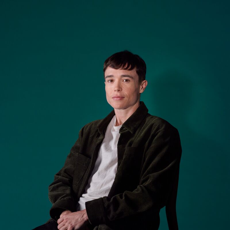 Elliot is pictured seated with his hands clasped and resting on his lap. He has cropped dark hair and wears a white t-shirt under a black jacket with black jeans.
