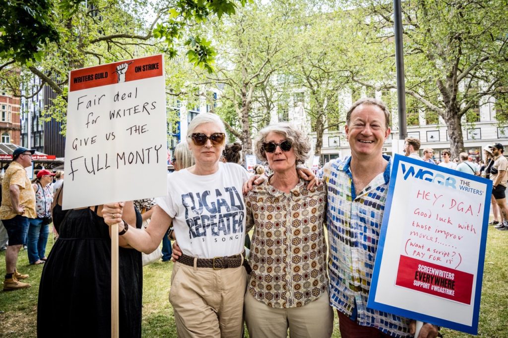 'The Full Monty' writers Alice Nutter and Simon Beaufoy stand with 'Misbehaviour' writer Gaby Chiappe between them at the Writers' Guild of Great Britain protest in solidarity with Writers Guild of America Strikes. Nutter is wearing a white t-shirt and light brown trousers, Chiappe wears a brown printed shirt and light brown trousers, Beaufoy wears a multi-coloured shirt. Nutter is holding a sign reading 'Fair deal for writers. Give us the Full Monty' while Beauty's sign reads 'Hey, DGA! Good luck with those improv movies...Want a script? Pay for it'. Protesters are visible in the background.
