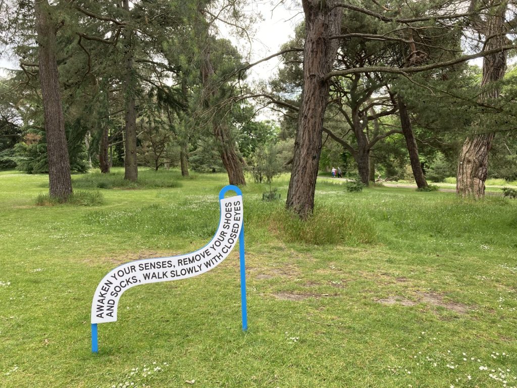 Artwork reading: Awaken your senses, remove your shoes and socks, walk slowly with closed eyes. Small sign in front of trees.
