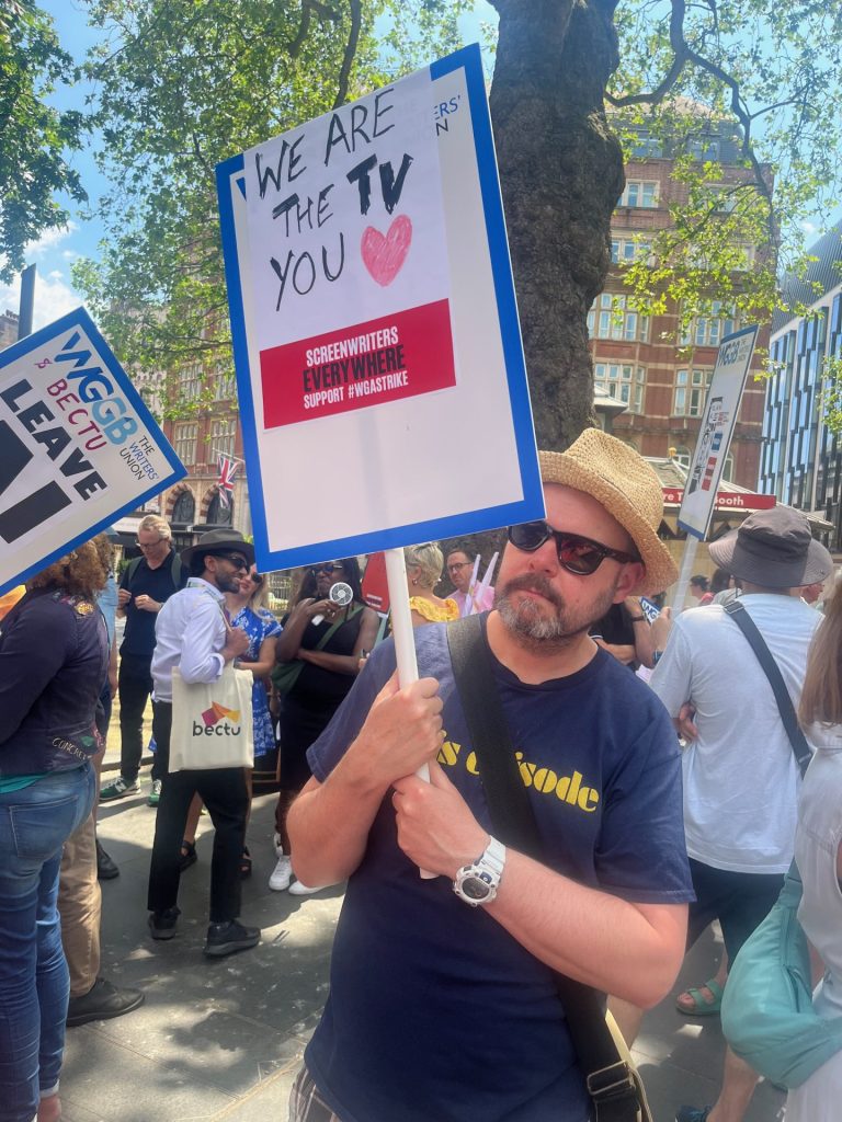 Writer Philip Lawrence at the Writers' Guild of Great Britain protest in solidarity with Writers Guild of America Strikes. He is holding a sign reading 'We are the TV you love' and wearing a navy-blue t-shirt, sunglasses and a straw hat.