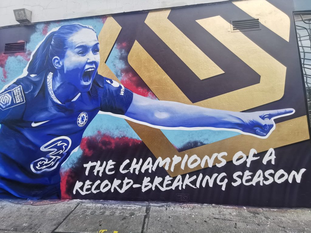 A picture of the mural on Portobello Road of Guro Reiten. Reiten has her arms outstretched in celebration. A line reading “The champions of a record-breaking season” is at the bottom of the mural, while the WSL logo in gold is part of the background.