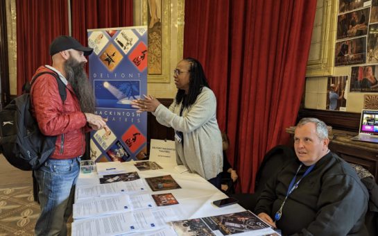 An attendee at the job fair stands in conversation with a representative from Delfont Mackintosh theatres, either side of a table, with a third person sat in the foreground.