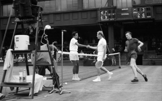 An archive image of two Wimbledon men's tennis players meeting at the net after a match, with Sam Hill, the ball boy, running round to collect a ball at the net.