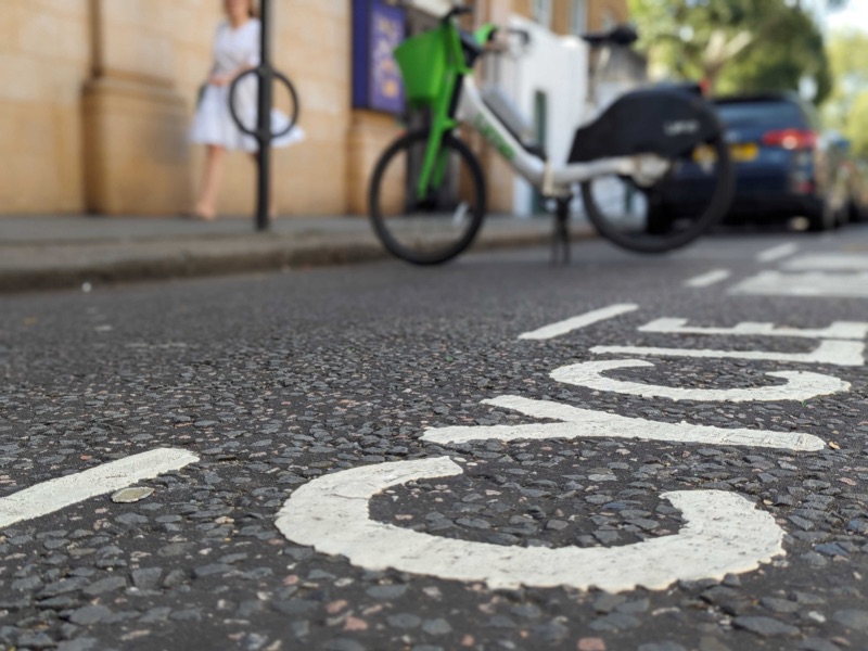 CYCLE in white paint on the road is the main focus. Blurred in the background is a Lime e-bike parked in the cycle parking spot