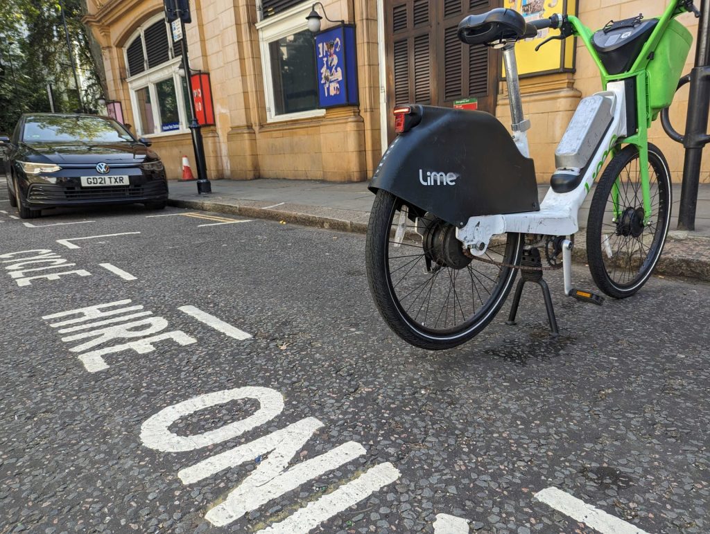 BIKE HIRE ONLY is written on the road in white paint. A lime rental e-bike sits alone in the park. Behind the park is a car