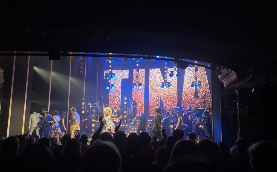 Karis Anderson as Tina Turner on stage surrounded by her backing band. A large light up sign saying "Tina" shines behind her in bright red.