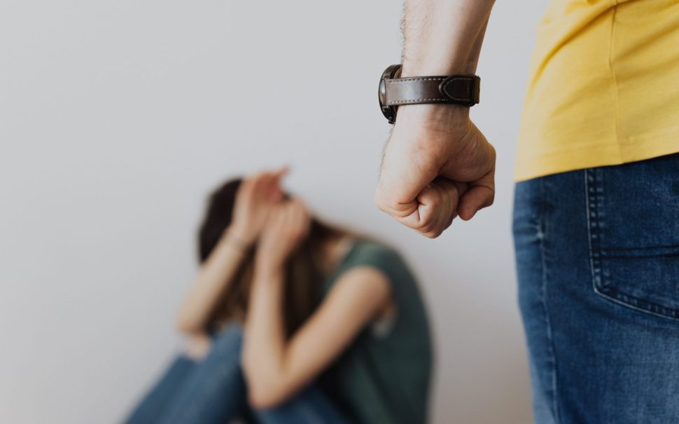 A man with a closed fist, stood over a woman sat on the floor with her hands over her face