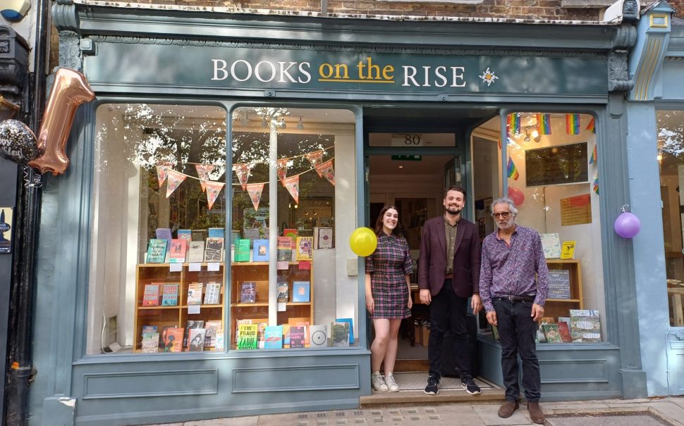 Two men and one woman standing outside Books on the Rise, a small bookshop, with balloons tied to the storefront