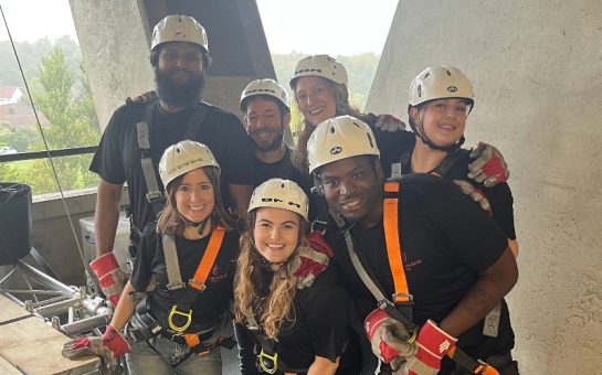 A group of fundraisers prepare to abseil from the top of the stadium. They are wearing hardhats and harnesses.