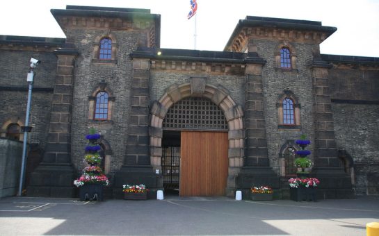 Wandsworth Prison, picture of front of the building, with wooden door and grey stone. Flowers in front of the building, sun shining