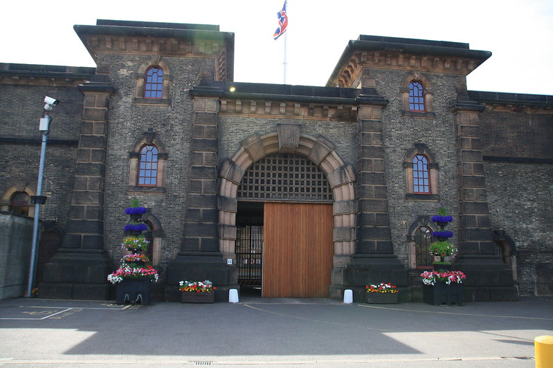 Wandsworth Prison, picture of front of the building, with wooden door and grey stone. Flowers in front of the building, sun shining