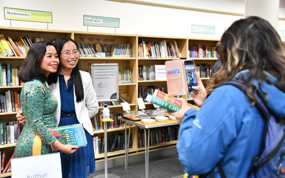 Author Nguyễn Phan Quế Mai with reader having their picture taken by another reader. They are holding the author's book and stood in the Merton Arts Space at Wimbledon Library.