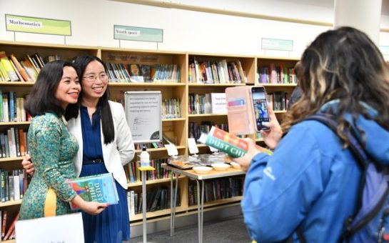 Author Nguyễn Phan Quế Mai with reader having their picture taken by another reader. They are holding the author's book and stood in the Merton Arts Space at Wimbledon Library.