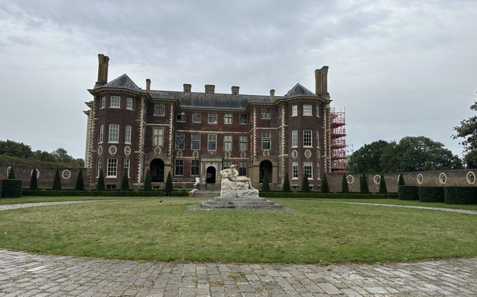 Picture of Ham House on a cloudy day, the building in red brick and has a statue infront of it. There is scaffolding to the right of the building.