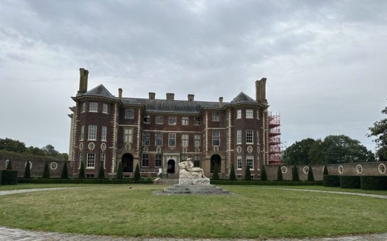 Picture of Ham House on a cloudy day, the building in red brick and has a statue infront of it. There is scaffolding to the right of the building.